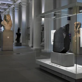 Pedra de Rosetta em uma vitrine retangular de vidro na galeria do Museu Britânico, com estátuas egípcias expostas em segundo plano.