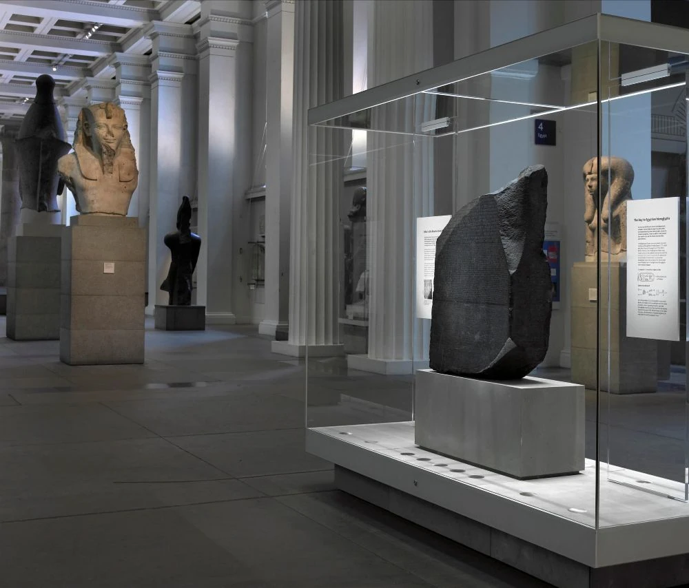 Pedra de Rosetta em uma vitrine retangular de vidro na galeria do Museu Britânico, com estátuas egípcias expostas em segundo plano.