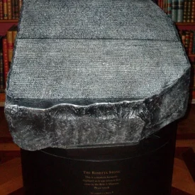 Réplica da Pedra de Rosetta em um pedestal redondo, com estantes de livros antigos ao fundo.