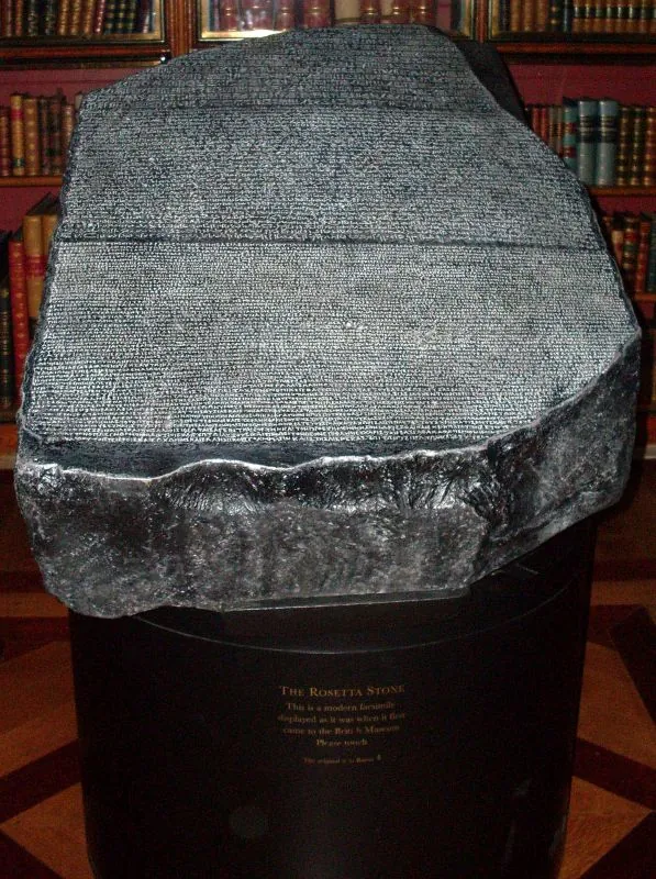 Réplica da Pedra de Rosetta em um pedestal redondo, com estantes de livros antigos ao fundo.